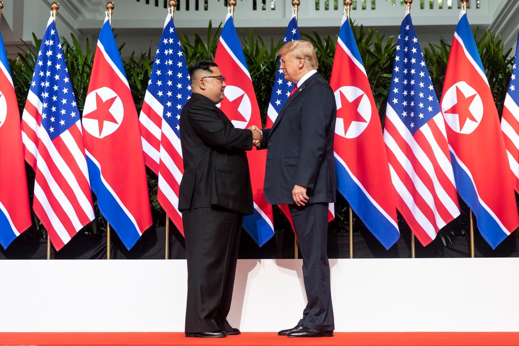 Kim Jong-un and Donald Trump shaking hands in front of a row of North Korean and American flags at the Capella Hotel, Singapore, June 2018.