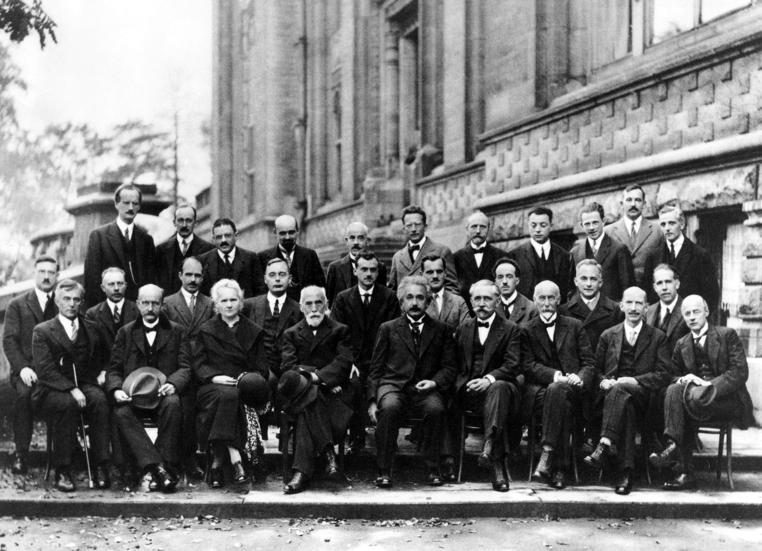 Twenty-nine physicists in formal dress arranged in three rows outside the Institut International de Physique in Brussels, 1927. Hendrik Lorentz sits at the centre of the front row with Marie Curie beside him.