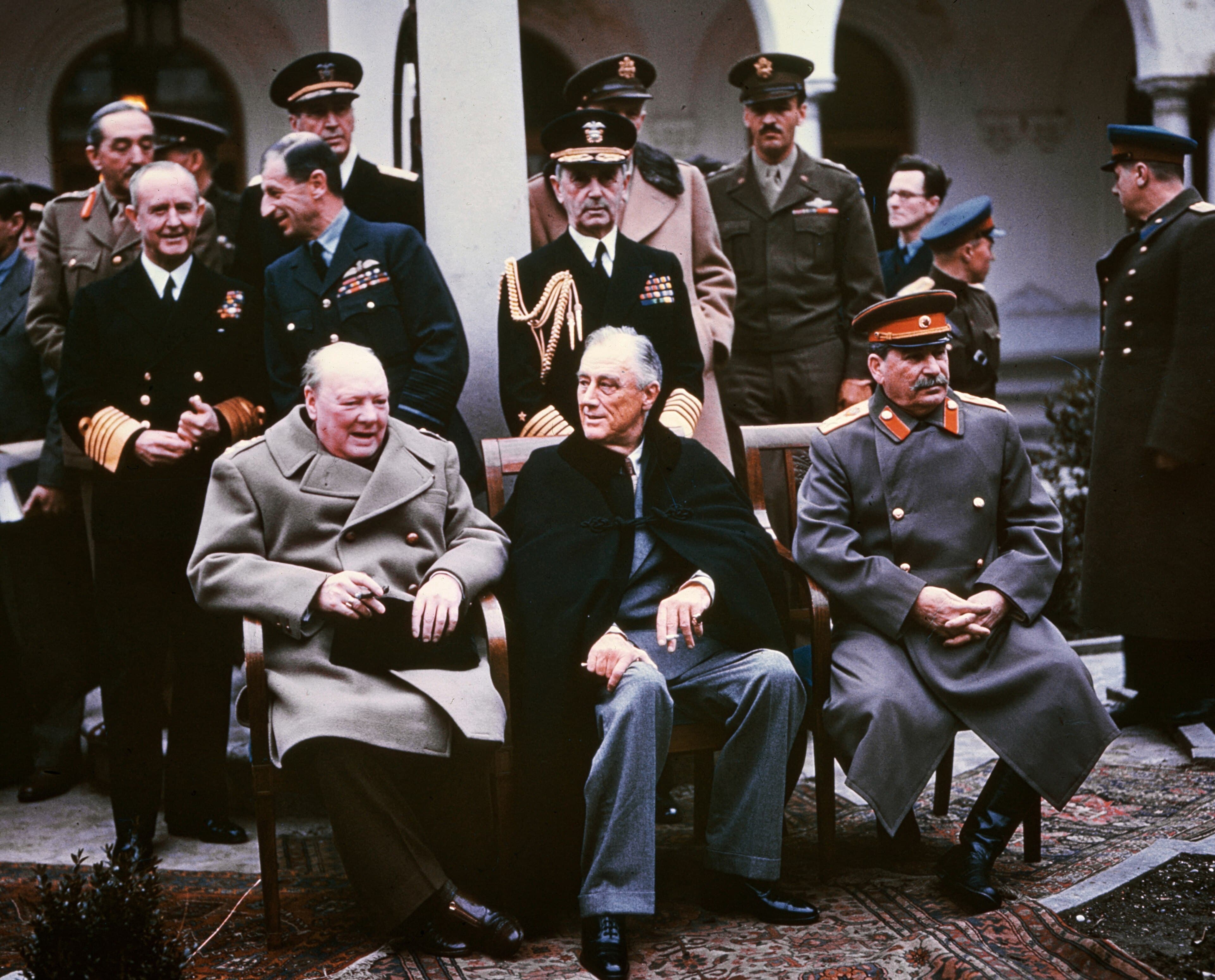 Winston Churchill, Franklin D. Roosevelt and Joseph Stalin seated together in the courtyard of Livadia Palace during the Yalta Conference, February 1945, with military and civilian staff standing behind them.